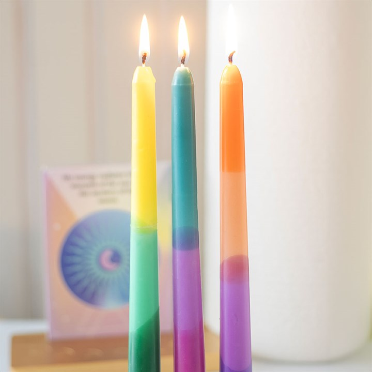Three colourful, vertically striped dipped taper candles—yellow-green, teal-purple, and orange-purple—stand lit side by side. A blurred book with a circular design and a white cylindrical object are in the background.