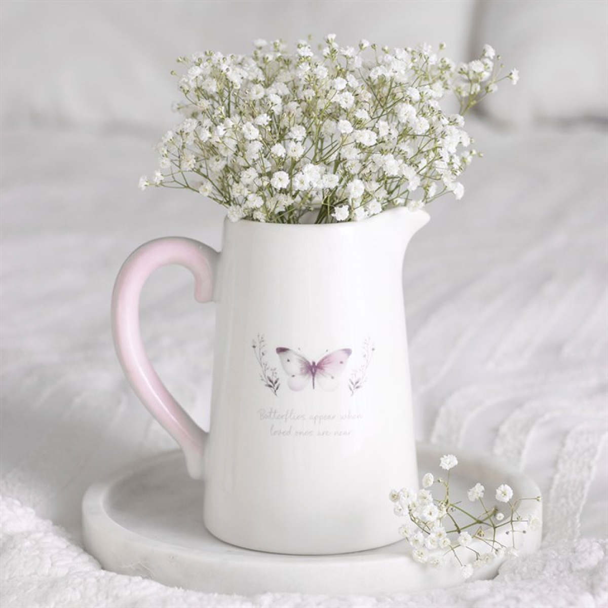 A decorative ceramic jug with a butterfly motif and pink handle holds delicate white baby's breath. Ideal as a memorial gift, it rests on a marble tray atop a white quilted surface.