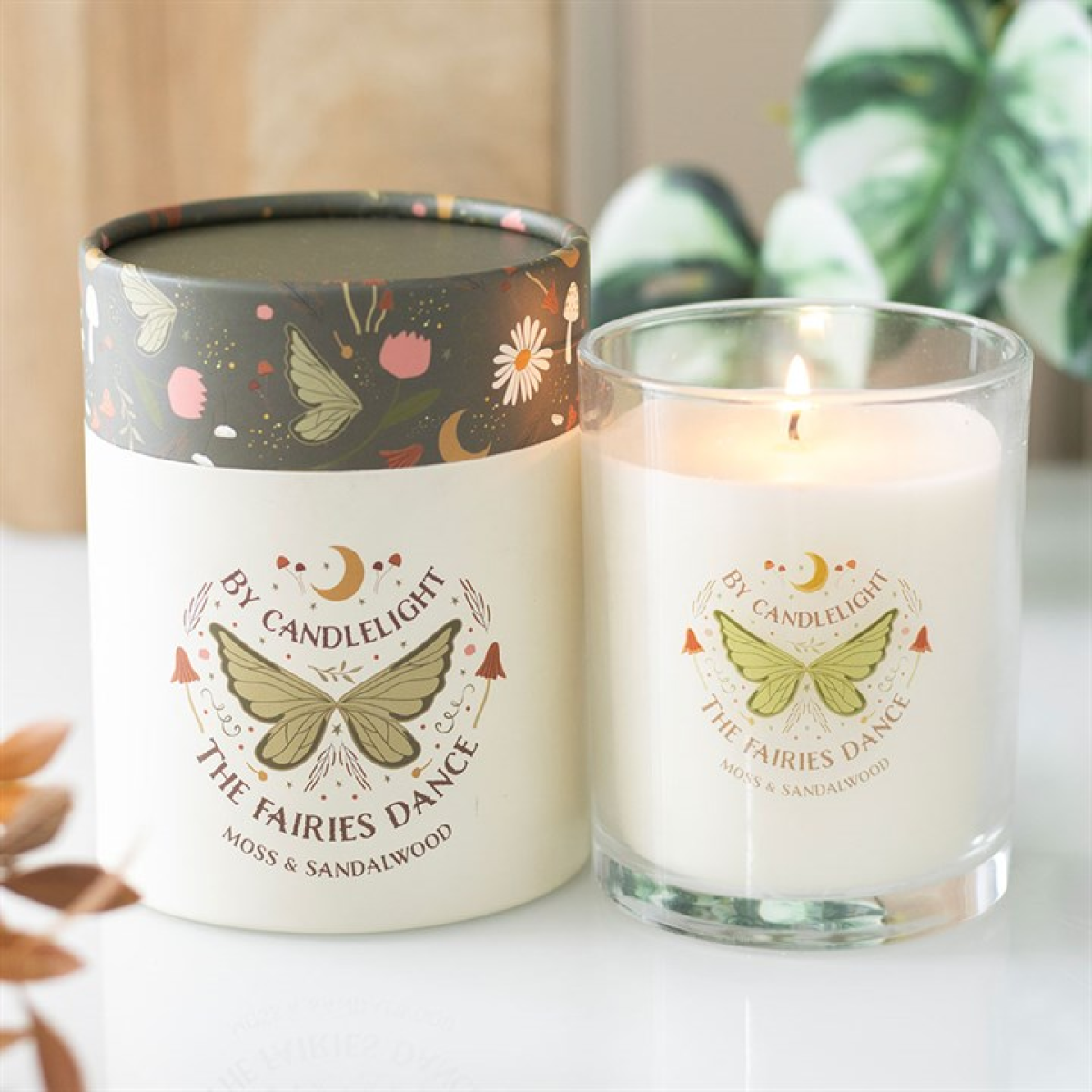 A lit Moss & Sandalwood candle in a fairy wing glass sits next to its cylindrical packaging, both decorated with whimsical butterfly and floral designs and the words: “By Candlelight, The Fairies Dance, Moss & Sandalwood.”.