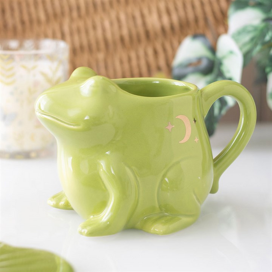A glossy green mug shaped like a frog, this quirky piece of homeware features a crescent moon and stars on its side. It rests on a white surface with a leafy plant and patterned glass in the background.