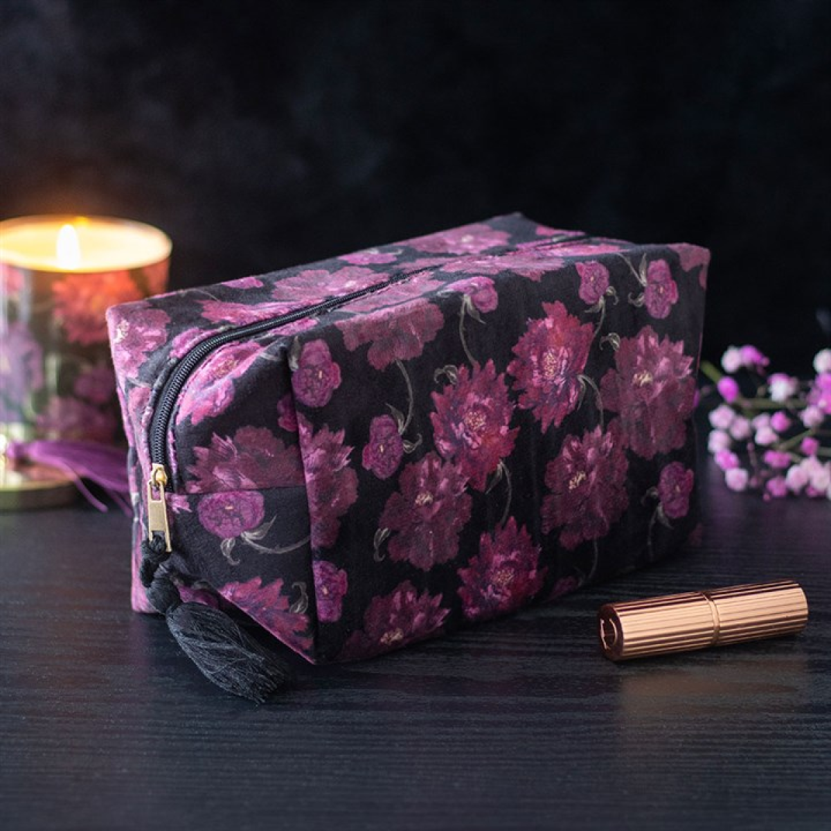 A rectangular make-up bag adorned with dark, gothic florals rests on a dark surface. Nearby, a lit candle, rose gold lipstick tube and purple flowers create a cosy, elegant scene.