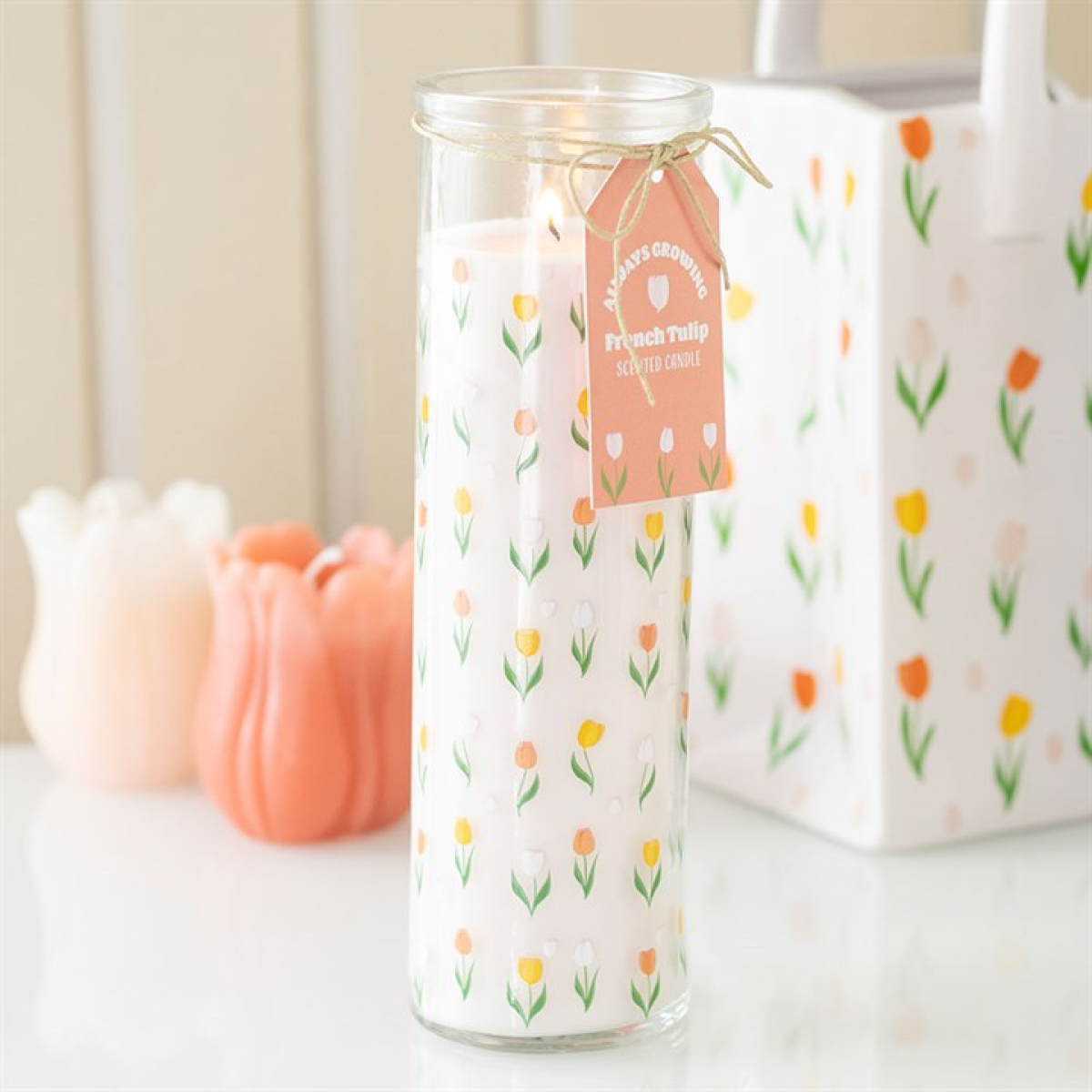 A tall tulip candle with a floral design and pastel tulips stands on a white surface. Its pink tag reads “French Tulip Scented Candle.” Candle holders and a coordinating gift bag are in the background.