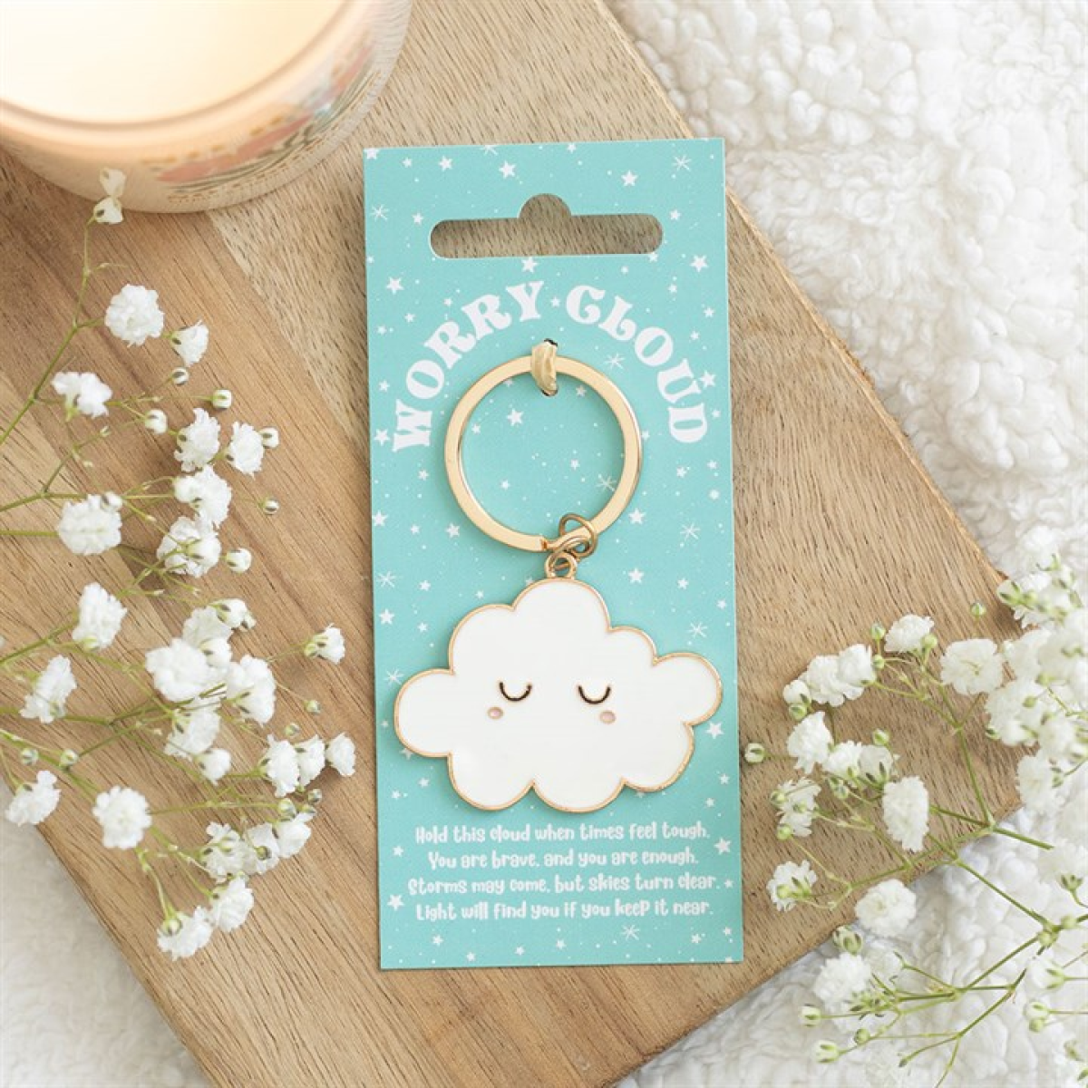 A charming enamel cloud keyring with a serene face rests on a blue card labelled Worry Cloud. This pocket-sized pick-me-up is displayed on a wooden tray with flowers, a candle, and a comforting message below.