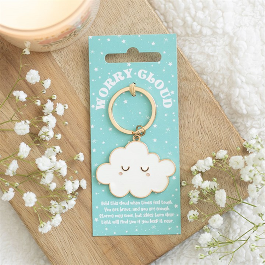 A charming enamel cloud keyring with a serene face rests on a blue card labelled Worry Cloud. This pocket-sized pick-me-up is displayed on a wooden tray with flowers, a candle, and a comforting message below.