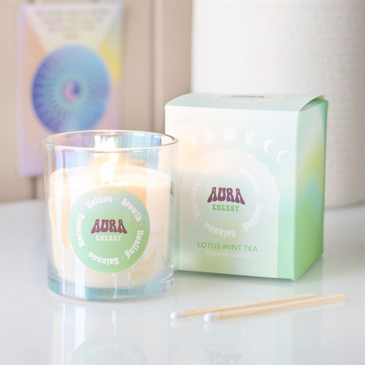 A lit iridescent glass candle labelled “Aura Cherry” sits next to a green box reading “Aura Cherry Lotus Mint Tea Scented Candle.” Two wooden matches and a blurred spiral card rest on the white surface nearby.