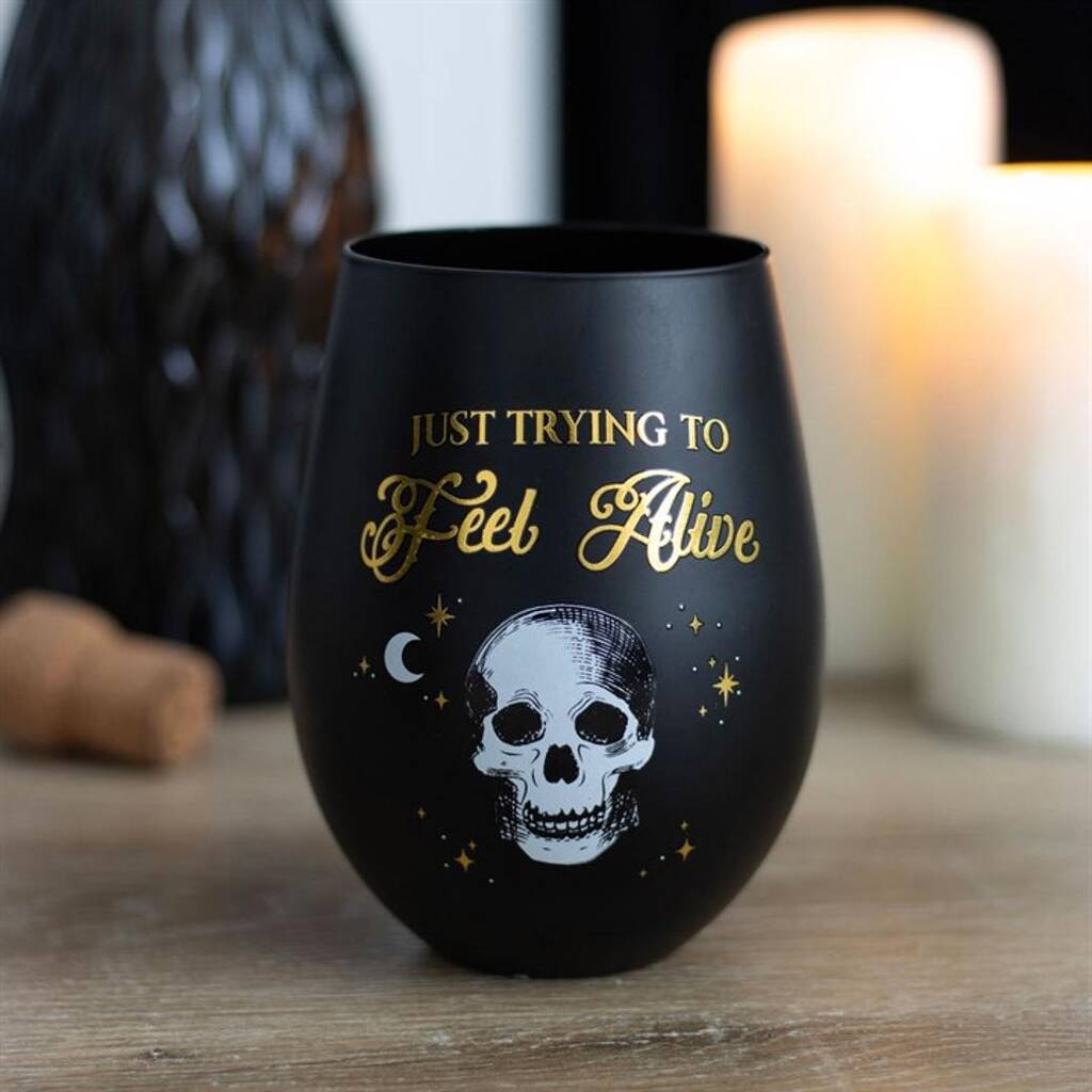 A black and gold wine glass bearing the words Just Trying To Feel Alive above a white skull, stars, and a crescent moon rests on a wooden surface, with lit candles softly blurred in the background.