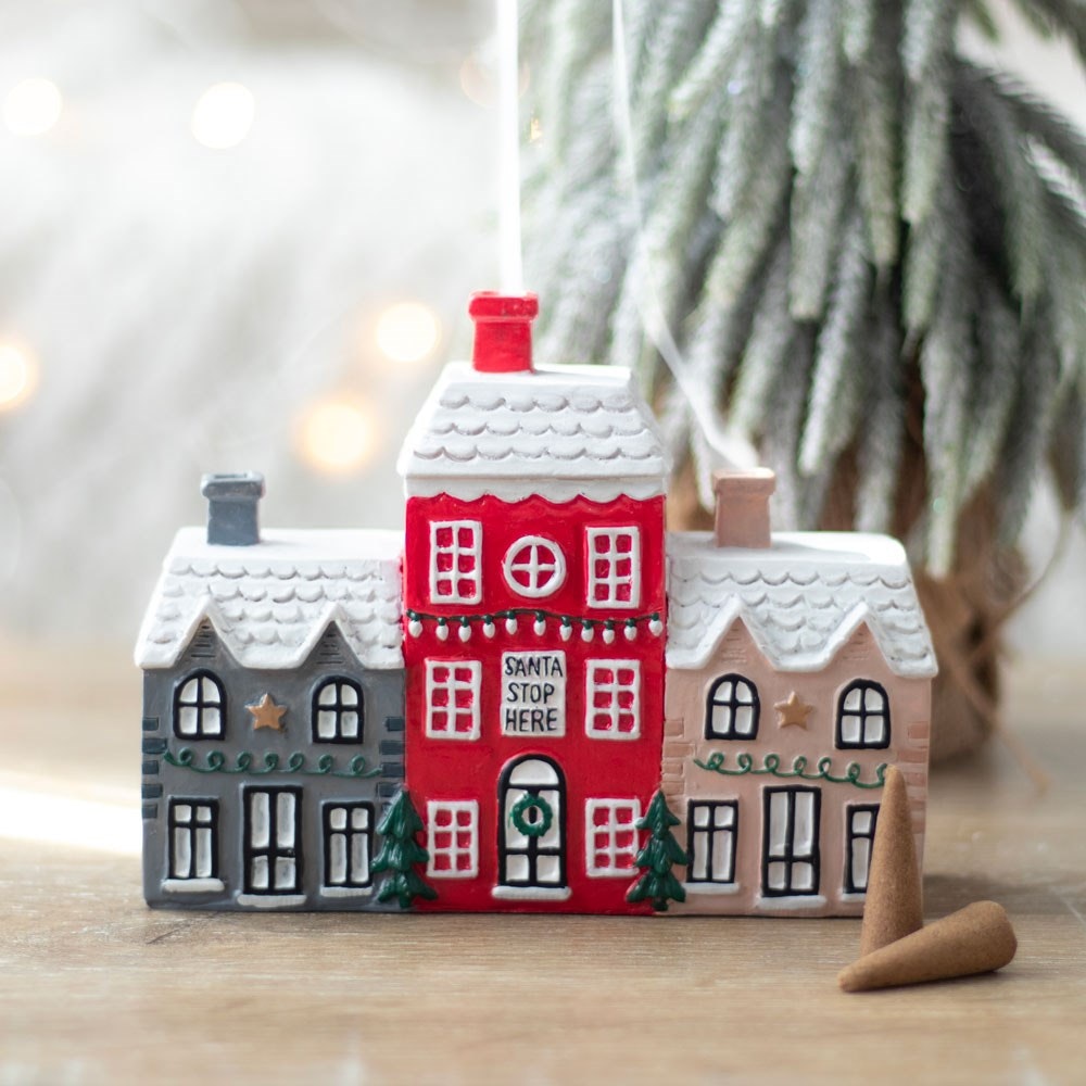 A festive ceramic ornament shaped like three snow-covered houses, with the middle red house bearing the words Santa Stop Here. This decorative Christmas piece includes small green trees, golden stars, and a blurred tree in the background.