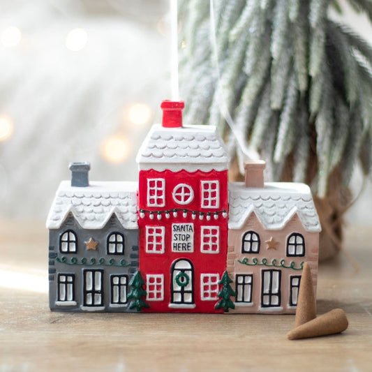 A festive ceramic ornament shaped like three snow-covered houses, with the middle red house bearing the words Santa Stop Here. This decorative Christmas piece includes small green trees, golden stars, and a blurred tree in the background.