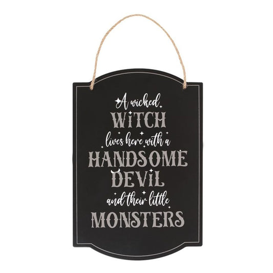 A black Halloween hanging sign with ornate white text reads: A wicked witch lives here with a handsome devil and their little monsters. The spooky décor features a jute rope for hanging and a decorative border that adds a whimsical touch.