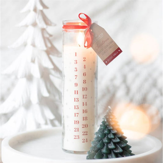 A white advent candle in a glass jar with red numbers 1 to 24, a red ribbon, and a tag. The lit, vanilla-scented candle rests on a white tray next to festive tree-shaped decorations.