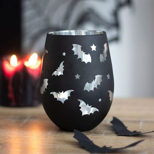 A black candle holder with bat and star cut-outs rests on a wooden surface next to a stemless glass decorated with bats. Two lit black candles cast a soft glow in the background, enhancing the Halloween atmosphere.
