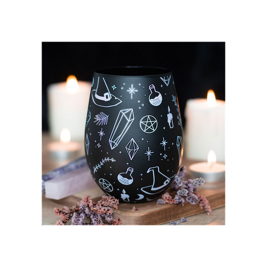 A black crystal glass decorated with purple and white mystical symbols—crystals, potion bottles, stars, pentagrams, and witch hats—sits on wood among dried flowers and flickering candles in a dreamy background.