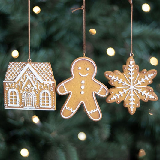 Three gingerbread ornaments—a house, a smiling gingerbread person, and a snowflake—hang as Christmas tree decorations on strings in front of a blurred evergreen with white festive lights in the background.