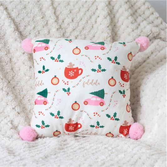 A pink pom pom cushion with festive Christmas prints—red mugs, pink cars with trees, candy canes, holly leaves, and baubles—rests on a cream textured blanket for charming festive home décor.