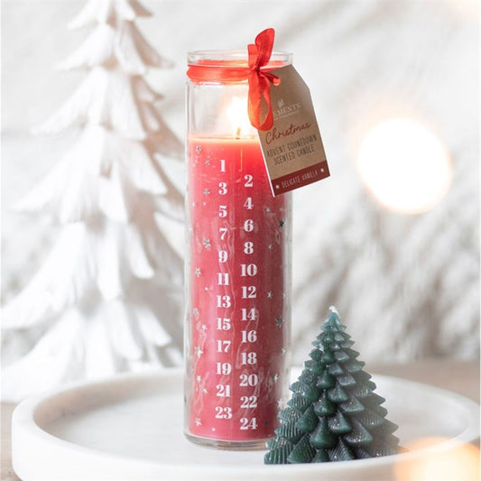 A tall red vegan paraffin wax advent candle, numbered in white from 1 to 24, burns inside a clear glass jar adorned with a red ribbon and tag. A green wax tree and a white decorative tree sit nearby on a white tray.
