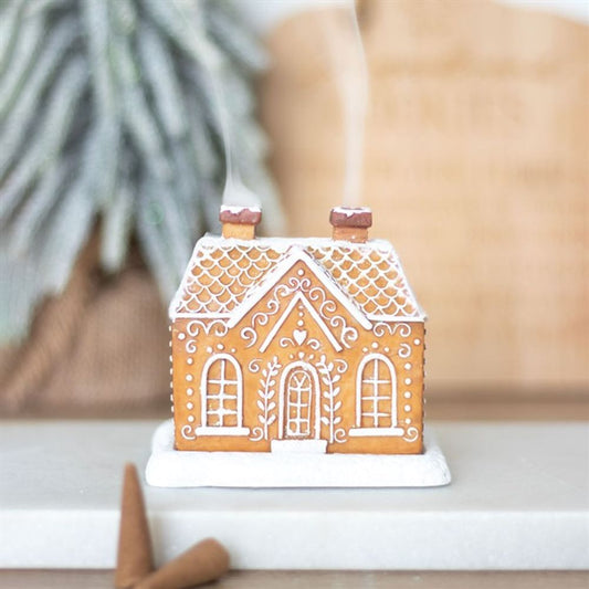 A gingerbread house with white icing details and two chimneys sits on a white surface. Light smoke, as from an incense cone burner, rises gently while cinnamon sticks rest in front—ideal for Christmas décor.