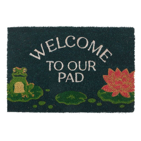 A welcome doormat with the text WELCOME TO OUR PAD features a charming frog design, complete with a cartoon frog on a lily pad to the left and a vibrant pink lotus flower to the right. Perfect for indoor use, its dark green background beautifully complements shades of green, pink, and white.