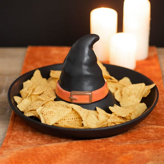 A black Halloween serving tray filled with assorted crisps surrounds a witch’s hat chip and dip platter with an orange band. Three lit pillar candles glow in the background on an orange cloth.