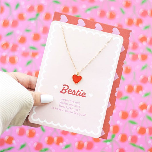 A hand with white-painted nails holds a card presenting a Bestie necklace, featuring a gold chain and red heart pendant. The card, perfect as a Valentines gift, reads Bestie alongside a friendship poem. A pink background with red cherry patterns and scalloped edges adds charm.