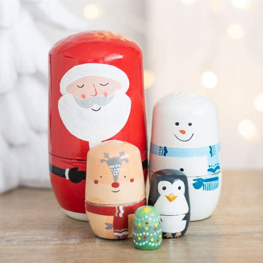 Four hand-painted Christmas decoration nesting dolls—Father Christmas, a snowman, reindeer, penguin, and a green Christmas tree—rest on a wooden surface with blurred white lights in the background.