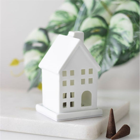 A small white ceramic incense holder shaped like a house with square windows rests on a white surface, with two brown incense cones nearby. In the background, green and white leaves are gently blurred.