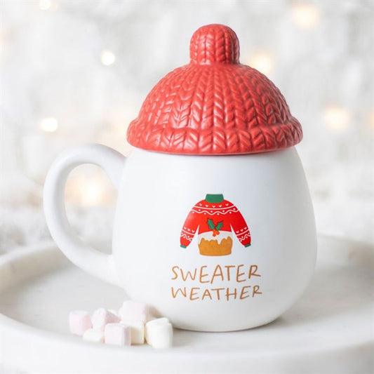This Christmas mug displays a red jumper illustration and the words SWEATER WEATHER. The novelty mug has a red knit-patterned lid resembling a beanie, with mini marshmallows nearby on the white surface.