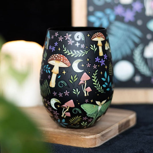 A black luna moth glass tumbler adorned with colourful illustrations of mushrooms, leaves, stars, moons, and flowers sits on a wooden surface, with a blurred candle and art print in the background.