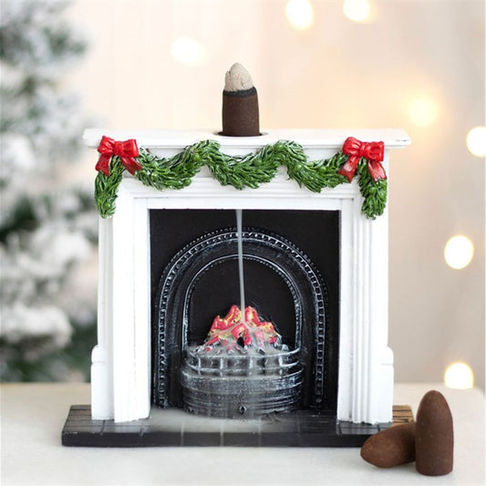 A miniature white Christmas fireplace decorated with festive garlands and red bows holds a backflow incense burner; smoke cascades down like fire and mist, set against a blurred, festive backdrop.