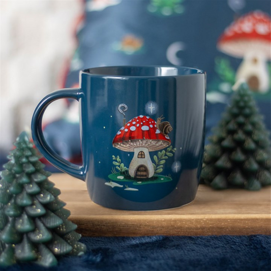 A navy-blue mushroom house mug featuring a red-capped mushroom home, green door, and snail rests on a wooden tray, flanked by pine tree candles. A matching cushion is visible in the blurred background.