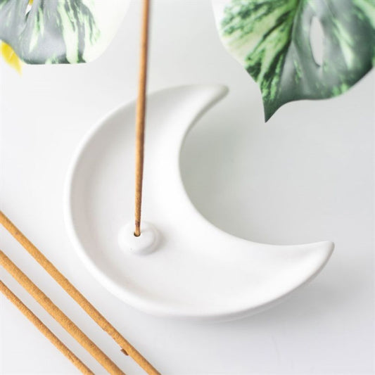 A contemporary ceramic incense holder shaped like a crescent moon holds a burning incense stick. Three unlit sticks lie nearby, with green leaves bordering the white background.