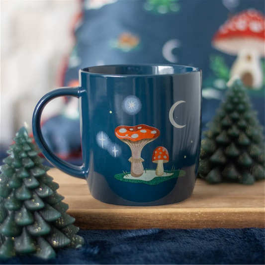 A navy blue mug adorned with red and white mushrooms, stars, and a crescent moon rests on a wooden surface between two small evergreen tree ornaments, with a matching cushion in the background for a magical toadstool mug feel.