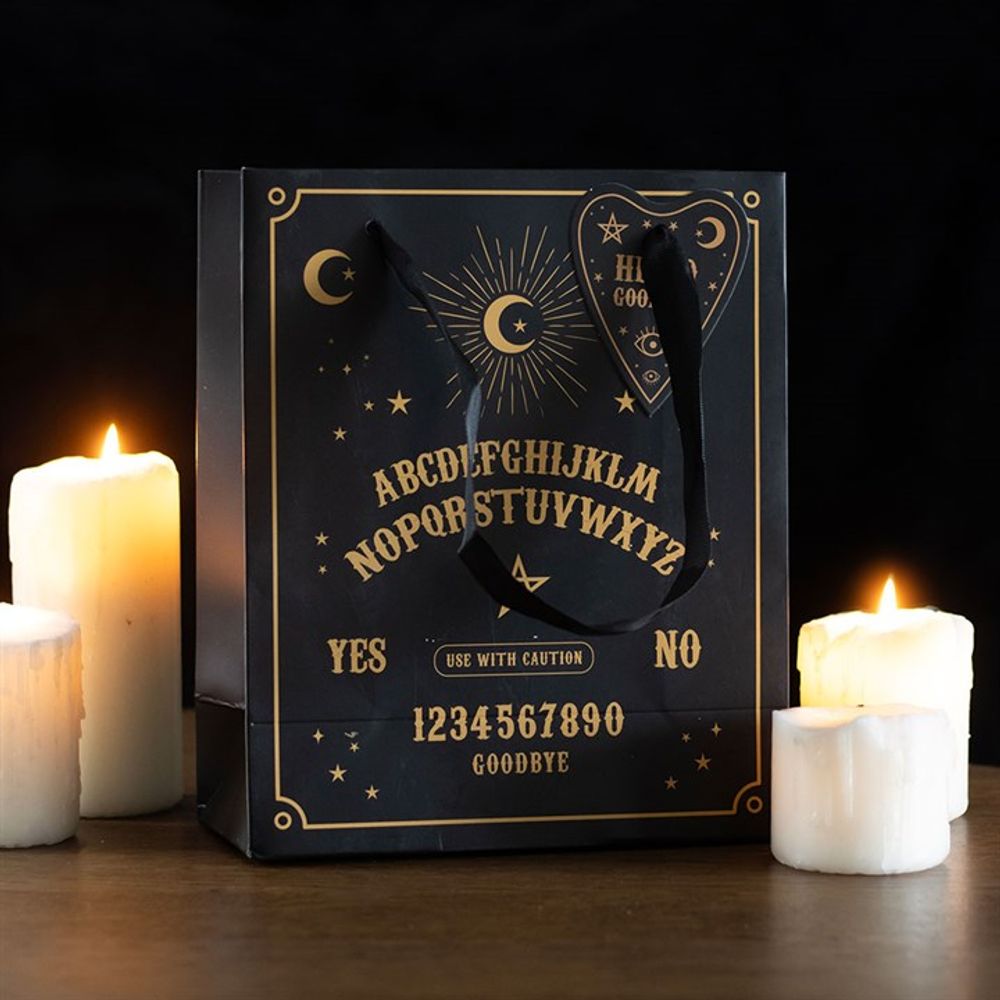 A black paper bag features a premium finish, adorned with ouija board elements in gold—including the alphabet, numbers, and the words YES, NO, GOODBYE. Black handles complement the design. Four lit white pillar candles surround this striking printed gift bag against a dark background.