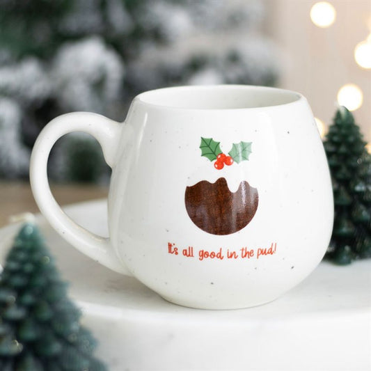 A festive mug styled as a Christmas pudding, adorned with holly details and red lettering saying It’s all good in the pud!—ideal next to pine decorations and a snowy, softly blurred festive background.