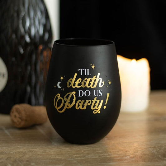 A black and gold wine glass with gold lettering reading “‘Til Death Do Us Party!” rests on a wooden surface beside a cork, a lit white candle, and a textured black bottle in the background.