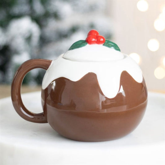 A festive piece of drinkware, this Christmas pudding mug has a handle, white icing detail, and a lid adorned with red berries and holly leaves. The blurred background glimmers with warm Christmas lights.