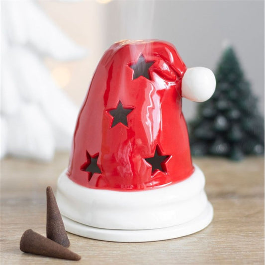 A red ceramic Christmas incense burner shaped like a Santa hat with star-shaped cut-outs rests on a white base. Smoke drifts from the top, while two brown incense cones lie close by. Blurred festive décor completes the scene.