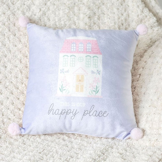 A soft white decorative cushion with a pastel blue and pink design of a house featuring pink windows and a roof. Below, the text reads This is our happy place. Light pink pom-poms adorn each corner, resting elegantly on a textured, creamy-white blanket—a cosy touch for any home décor.