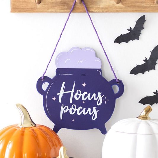 A purple Hocus Pocus cauldron hanging sign sits above ceramic pumpkins, while black bats on the white wall complete this charming Halloween décor scene.