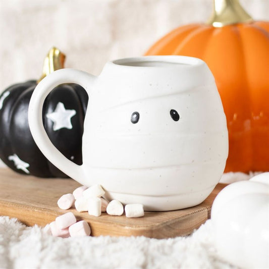 A spooky mummy mug with two black eyes sits on a wooden board, surrounded by mini marshmallows. Black and orange pumpkins are in the background on a soft, white textured surface.