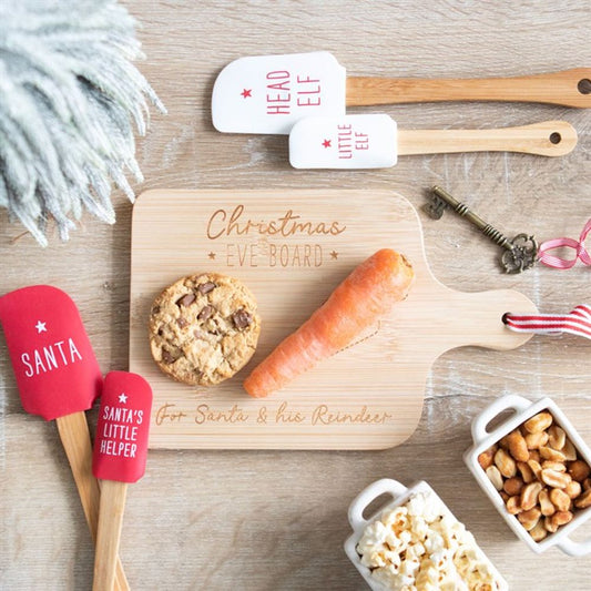 A festive Christmas Eve serving board labelled for Father Christmas & his Reindeer holds Father Christmas’s biscuits and a carrot. Surrounding it are red and white spatulas, popcorn, and nuts on a light wooden surface.