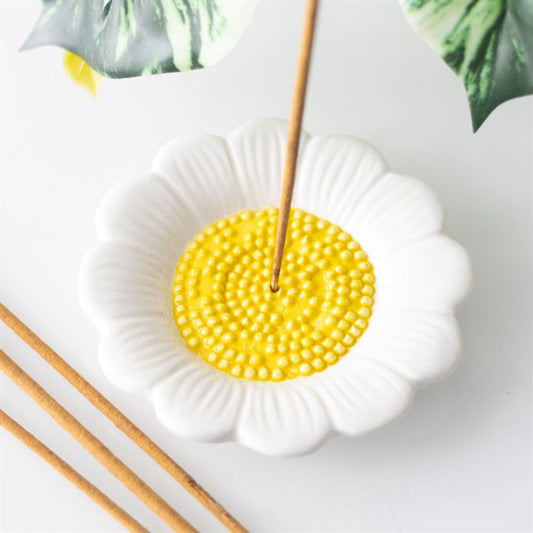 A white ceramic incense holder, designed like a daisy with a textured yellow centre, cradles a single lit incense stick while two unlit sticks rest nearby. An elegant ash catcher design is enhanced by green leaves partially visible on the pristine white surface.