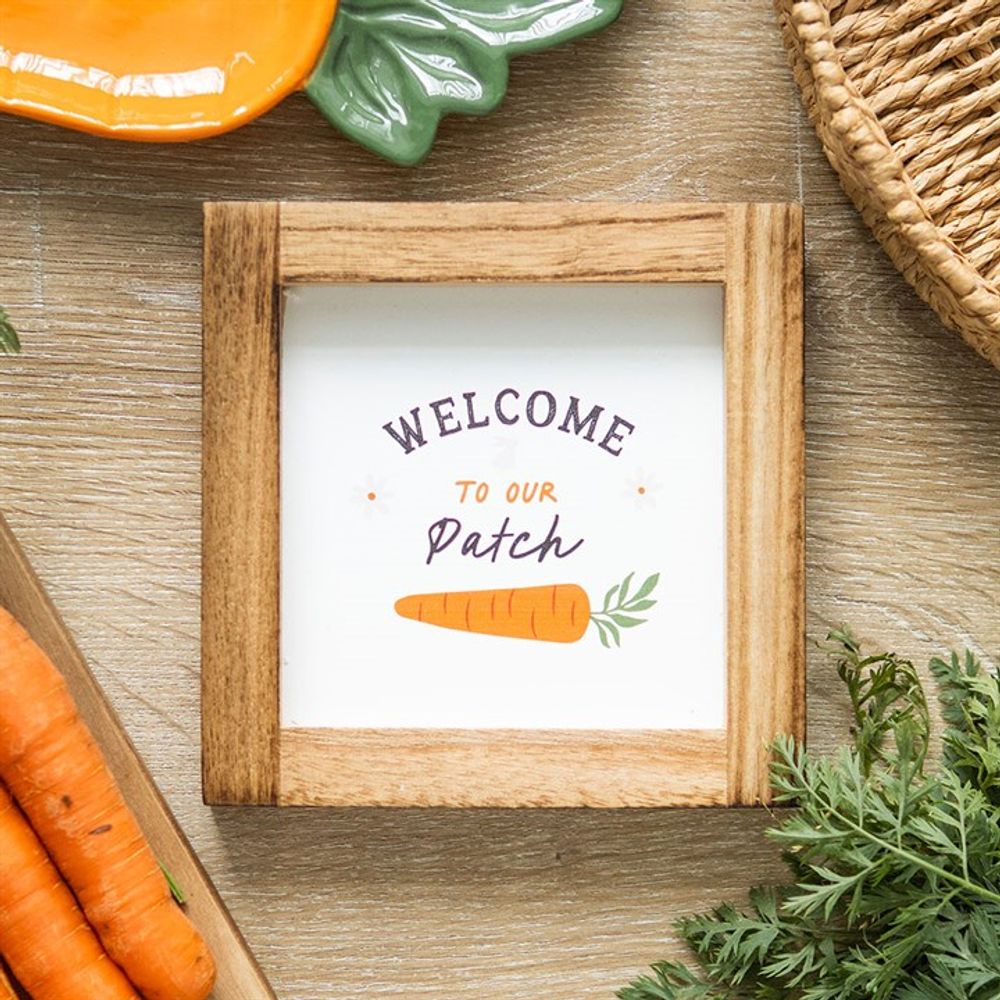 A square wooden frame sign on a table displays Welcome to our Patch above an illustration featuring carrot details. Nearby, a woven basket, a green ceramic dish, and several fresh carrots with leafy tops create a charming arrangement perfect for any bunnys delight on the wooden surface.