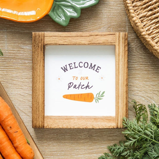 A square wooden frame sign on a table displays Welcome to our Patch above an illustration featuring carrot details. Nearby, a woven basket, a green ceramic dish, and several fresh carrots with leafy tops create a charming arrangement perfect for any bunnys delight on the wooden surface.