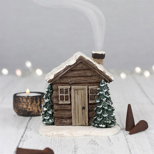 A small, rustic log cabin incense burner with a snow-covered roof and two snow-dusted pine trees sits on a light wooden surface. Thin smoke rises from its chimney, adding to the cosy winter décor aglow with soft fairy lights.