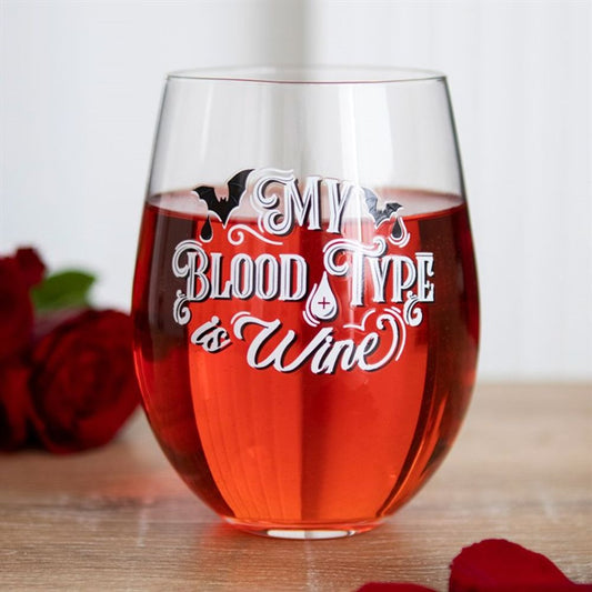 A stemless wine glass filled with red wine, featuring white gothic text reading, My blood type is wine, along with a bat motif and a drop of blood. Red rose petals and roses lie on the wooden surface beside the glass.