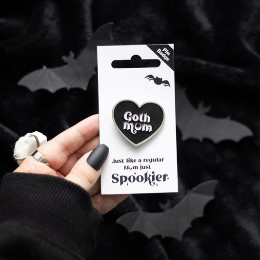 A hand with black matte nails and silver rings holds a black heart pin reading “Goth Mum.” The backing card says, “Just like a regular Mum, only spookier,” with bat silhouettes on a black velvet background.