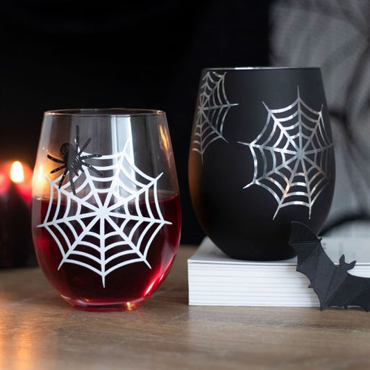 Two stemless wine glasses with white spiderweb designs—one clear with red liquid and a spider, the other black—sit next to a paper bat and lit candle, creating a spooky Halloween-themed scene.