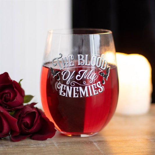 A gothic wine glass filled with red liquid, labelled “The Blood of My Enemies,” rests on a wooden surface next to two red roses and a lit candle, set against striking dark and white backgrounds.
