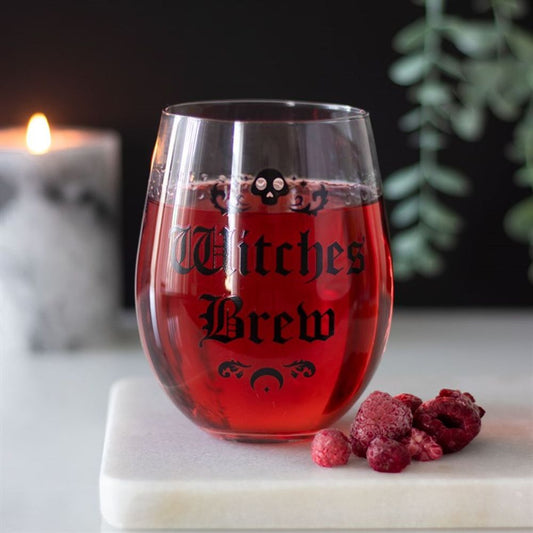 A stemless magic potion glass featuring Witches Brew and skull motifs contains a red drink. Frozen berries sit on a white board, with a glowing candle and green plant gently blurred in the background.